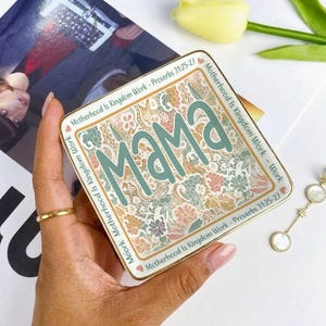 Puede incluir: Una bandeja cuadrada de cerámica con borde dorado. La bandeja presenta la palabra "mama" en escritura turquesa sobre un estampado floral. El texto "Motherhood Is Kingdom Work - Proverbs 31:25-27" está impreso alrededor del borde.
