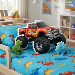 May include: A red and yellow monster truck toy with the name "Jackson" on the side, parked on a bed with a blue dinosaur-themed comforter. Two plush dinosaur toys are on the bed. The truck has large black tires and a silver front bumper.