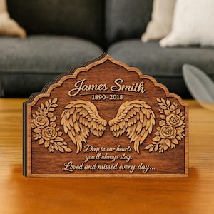 May include: A wooden memorial plaque with the name "James Smith" and dates 1890-2018. The plaque features carved angel wings, floral accents, and the inscription "Deep in our hearts you'll always stay. Loved and missed every day..."