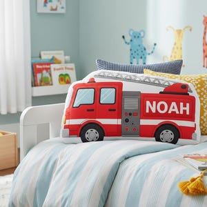 May include: A red fire truck shaped pillow with the name "NOAH" in white letters. The pillow has black wheels, a silver ladder, and is on a bed with a blue and white striped comforter. Decorative pillows are in the background.