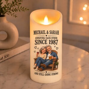 Op de afbeelding: Een cilindervormige, crèmekleurige led-kaars met een bedrukte illustratie van een koppel dat op een houten stoel zit. De tekst luidt: MICHAEL & SARAH, ANNOYING EACH OTHER SINCE 1987, AND STILL GOING STRONG. De kaars geeft een warm licht af.