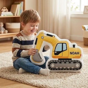 Op de afbeelding: Een geel en wit kussen in de vorm van een graafmachine met de naam "NOAH" aan de zijkant. Het kussen heeft zwarte en grijze details, waaronder de rupsbanden en de arm. Een kind houdt het kussen vast.