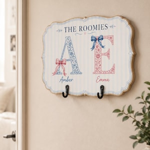 May include: A rectangular wall-mounted sign with scalloped edges featuring the text THE ROOMIES, the letters A and E decorated with floral patterns, a pink bow on the A, a blue bow on the E, the names Amber and Emma, and two black metal hooks.