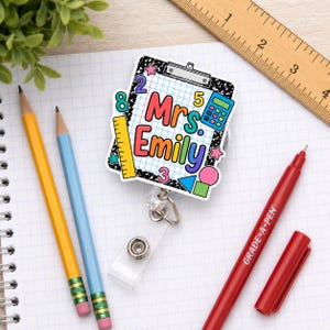 Peut inclure: Un enrouleur de badge avec un motif de presse-papiers portant le texte Mrs. Emily, entouré de graphismes scolaires colorés incluant une calculatrice, une règle et des chiffres sur un fond moucheté noir et blanc, placé près d'une règle en bois.