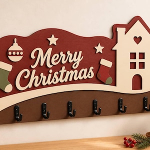 May include: A wall-mounted wooden key holder featuring a dark red background with the text Merry Christmas in cream lettering. The design includes a cream-colored house, two stockings, and two stars. It has six black metal hooks along the bottom.