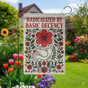 Op de afbeelding: Een tuinvaandel met de tekst "RADICALIZED BY BASIC DECENCY" in bordeauxrood. De vlag heeft een rode bloem, een witte duif en bloemmotieven op een beige achtergrond. De vlag wordt getoond in een tuin met bloemen.
