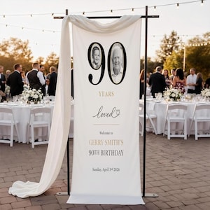 May include: A white banner with black text and two photos, celebrating a 90th birthday. The banner reads "90 Years Loved" and welcomes guests to Gerry Smith's 90th Birthday on April 3rd, 2026. The banner is displayed at an outdoor event.