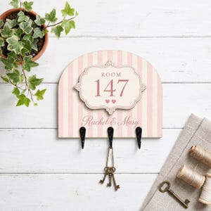 May include: A semi-circular wall-mounted key holder with a light pink and white striped pattern. It features a central plaque labeled ROOM 147 with two small hearts and the names Rachel & Mary. Three black metal hooks hold a set of metal keys.