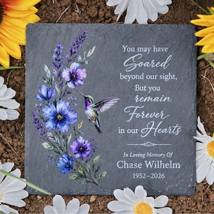 May include: A square, gray memorial stone with floral and hummingbird artwork. The stone features blue and purple flowers, a hummingbird, and the inscription "You may have Soared...Forever in our Hearts." It also includes "In Loving Memory Of Chase Wilhelm 1952-2026."