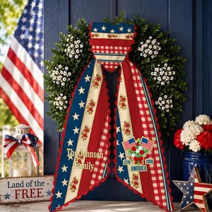 Peut inclure: Une couronne décorative avec un ruban patriotique aux motifs rouges, blancs et bleus, étoiles et fleurs. Le ruban affiche le texte The Johnson Family, 1776, 2026 et AMERICA, accompagné d'une illustration de deux drapeaux américains.