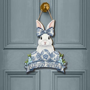 May include: A wooden Easter door hanger featuring a white bunny with a blue patterned bow and eggs. The banner below reads "Happy Easter" in blue script. The door hanger is attached to a gray door with a gold door knocker.