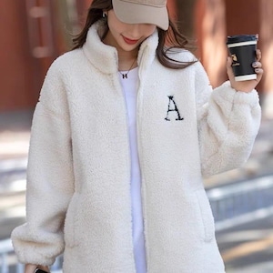 May include: Cream-colored, fluffy fleece jacket with a high collar and a black embroidered "A" on the chest. The jacket has a full-length zipper and two front pockets. A beige baseball cap is worn.