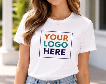 May include: White t-shirt with the text "YOUR LOGO HERE" in a square frame. The text is in orange, teal, and navy blue. The model is wearing blue jeans. The background is blurred, with greenery and a white building.