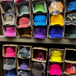 May include: A variety of colorful baseball caps are neatly arranged in cardboard boxes on shelves. The caps come in various colors, including pink, green, yellow, blue, and purple. Some are individually wrapped in clear plastic.