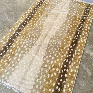 May include: Rectangular rug with a spotted pattern in shades of brown, tan, and cream. The rug has a fringed edge and a striped design. The rug is made of woven material.