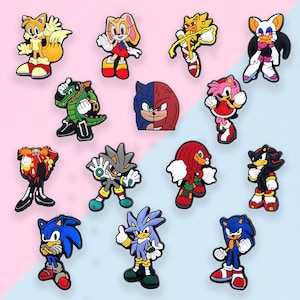 May include: A collection of colorful cartoon character charms, featuring various characters from the Sonic the Hedgehog franchise. The charms are in various poses and colors, including blue, red, yellow, and green, against a pink and blue background.