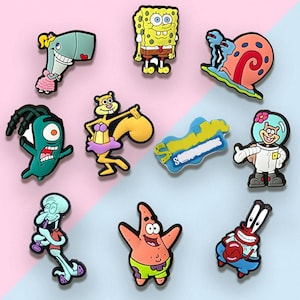 May include: A collection of colorful cartoon character charms, including SpongeBob SquarePants, Patrick Star, and Squidward. The charms are made of a glossy material and feature vibrant colors and detailed designs. The background is a light pink and blue.
