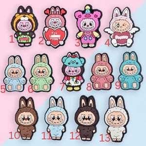 Puede incluir: Una colecci&oacute;n de trece amuletos de personajes de conejos de dibujos animados coloridos. Cada amuleto presenta un dise&ntilde;o &uacute;nico, con variaciones en color, expresiones faciales y accesorios. Los amuletos est&aacute;n numerados del 1 al 13 y est&aacute;n sobre un fondo azul y rosa.