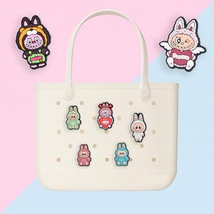 Puede incluir: Bolso tote de color crema con asa, decorado con coloridos amuletos de conejitos de dibujos animados. Los amuletos son de varios colores, incluyendo verde, rosa y azul. El fondo est&aacute; dividido entre rosa y azul.