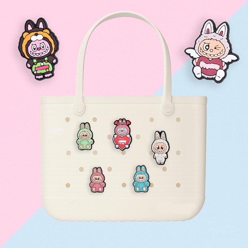 Puede incluir: Bolso tote de color crema con asa, decorado con coloridos amuletos de conejitos de dibujos animados. Los amuletos son de varios colores, incluyendo verde, rosa y azul. El fondo est&aacute; dividido entre rosa y azul.