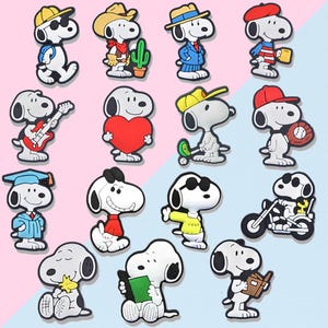 May include: A collection of Snoopy character charms in various poses and outfits. The charms feature Snoopy with a guitar, a heart, a graduation gown, sunglasses, and more. The background is a split of pink and light blue.