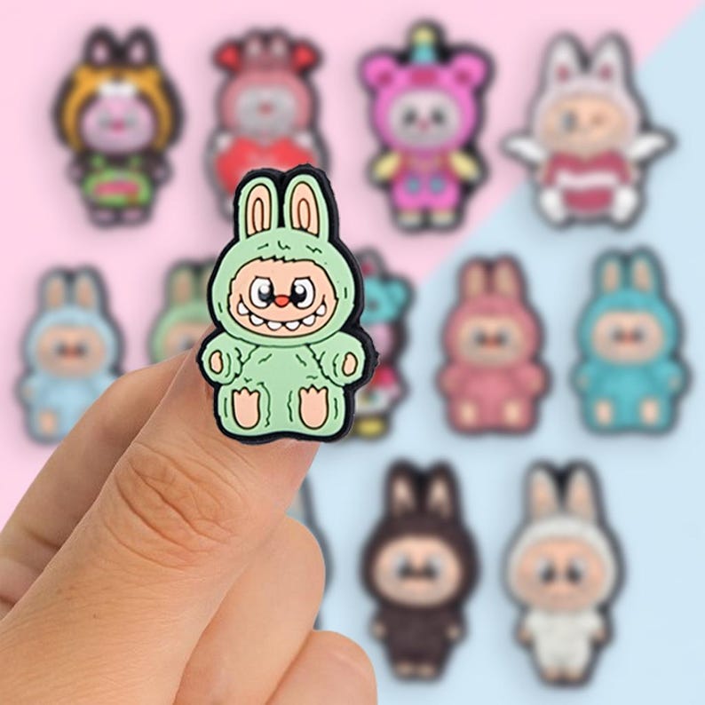 Puede incluir: Un amuleto verde con forma de conejito con una cara sonriente, rodeado de otros amuletos de dibujos animados coloridos. Los amuletos tienen varios dise&ntilde;os de animales y personajes, con un fondo borroso rosa y azul.