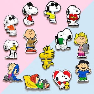 May include: A collection of colorful Snoopy and Peanuts character charms. The charms feature Snoopy in various poses, including wearing sunglasses, holding a heart, and reading a book. Other characters include Charlie Brown, Woodstock, and Lucy.