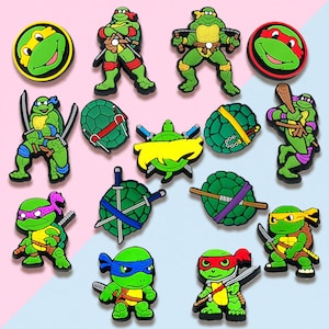May include: A collection of colorful Teenage Mutant Ninja Turtles character charms. The charms feature the turtles in various poses, with weapons, and their signature colored masks. The charms are green, red, blue, purple, and yellow.