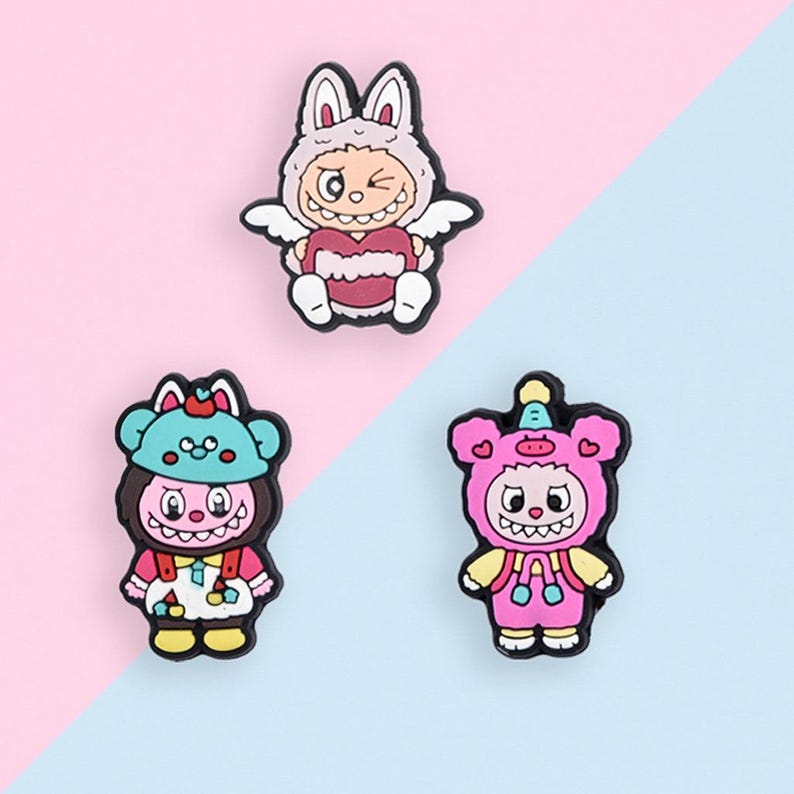 Puede incluir: Tres coloridos amuletos de personajes de dibujos animados. El amuleto superior es una figura con un disfraz de conejito con alas. El amuleto inferior izquierdo es una figura con sombrero y peto. El amuleto inferior derecho es una figura con un disfraz de oso.