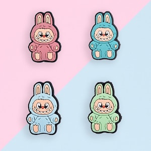 Puede incluir: Cuatro figuras de conejito de dibujos animados de colores en rosa, azul, azul claro y verde. Cada figura tiene una cara sonriente, orejas de conejo y un contorno negro. El fondo est&aacute; dividido diagonalmente, con secciones rosas y azul claro.