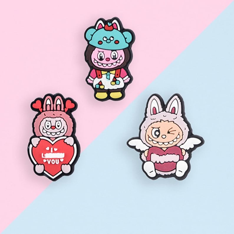 Puede incluir: Tres amuletos de personajes de dibujos animados coloridos. Un amuleto es un oso con un sombrero azul. Otro amuleto es un conejito que sostiene un coraz&oacute;n con el texto "I &hearts; YOU". El tercer amuleto es un conejito con alas que sostiene un coraz&oacute;n.