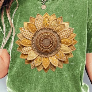 Pode incluir: Uma camiseta verde com um grande desenho de girassol. O girassol é feito de tecido amarelo e bege, com um centro texturizado. A camiseta tem uma aparência ligeiramente desbotada.