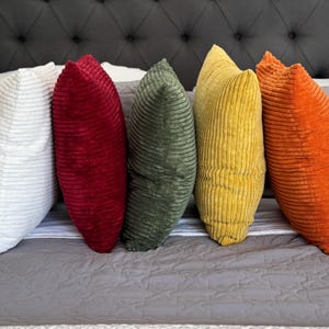 May include: Five corduroy throw pillows in white, red, green, yellow, and orange are arranged on a bed. The pillows are in a row, leaning against a gray tufted headboard and a gray quilted bedspread.