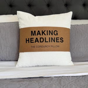 Könnte beinhalten: Ein weißes Cordkissen mit einem braunen Band mit der Aufschrift "MAKING HEADLINES" und "THE CORDUROY PILLOW" in schwarzer Schrift. Das Kissen liegt auf einem Bett mit grauer und weißer Bettwäsche.