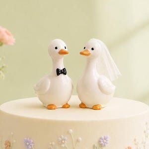 May include: Two white duck figurines placed on top of a round cake. The left duck wears a small black bow tie, and the right duck wears a white veil. Both ducks have orange beaks and feet. The cake is decorated with small, delicate floral patterns.
