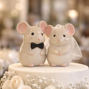 Può includere: Due statuette di topo in ceramica posizionate una accanto all'altra. Il topo a sinistra indossa un papillon nero. Il topo a destra indossa un velo di tulle bianco. Entrambi hanno corpi beige chiaro, orecchie rosa, occhi neri e naso rosa.