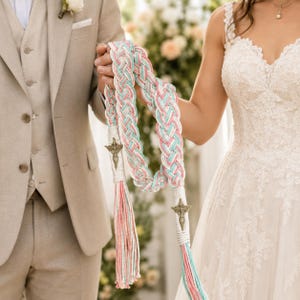 Puede incluir: Un cordón de boda trenzado en colores pastel rosa, azul y blanco. El cordón presenta cruces metálicas decorativas y borlas. El cordón es sostenido por una pareja, la novia viste un vestido de novia de encaje blanco y el novio un traje color beige.