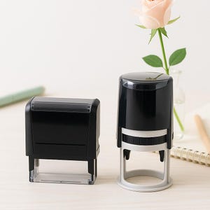 May include: Two black self-inking stamps, one rectangular and one round, are displayed on a light-colored surface. The rectangular stamp is on the left, and the round stamp is on the right. A light pink rose in a small glass vase is in the background.
