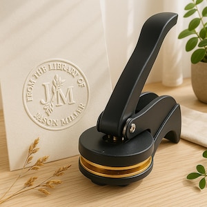 May include: A black and gold embosser with a long handle, next to a cream-coloured card with the embossed text "FROM THE LIBRARY OF JASON MILLER". The embosser has a circular base with gold accents.