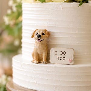 May include: A light brown Labrador retriever figurine sitting on a white tiered cake. Next to the dog is a small rectangular sign with the text I DO TOO and a tiny pink heart. The cake has a textured horizontal finish and white frosting.