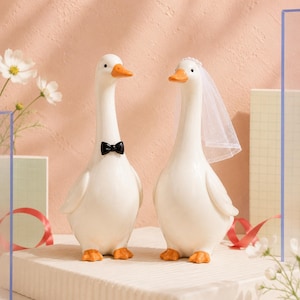 May include: Two white ceramic goose figurines standing side by side. The left goose wears a small black bow tie. The right goose wears a white sheer veil with small white beads along the top edge. Both geese have orange beaks and orange feet.