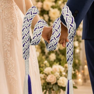 Puede incluir: Cordón de boda trenzado en blanco, gris y morado. El cordón es sostenido por dos personas, con un fondo floral. El cordón tiene una borla en el extremo con hebras blancas, grises y moradas. Un símbolo de unidad.