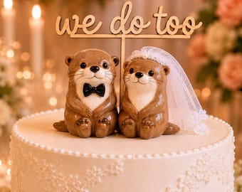 Può includere: Un topper per torta con due lontre in ceramica marrone sedute fianco a fianco. Una lontra indossa un papillon nero, l'altra un velo di pizzo bianco. Sopra di loro c'è un cartello in legno con la scritta "we do too" in carattere corsivo.