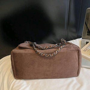 May include: A brown suede shoulder bag with a silver chain strap. The bag has a rectangular shape with a flap closure. The bag is on a white surface.