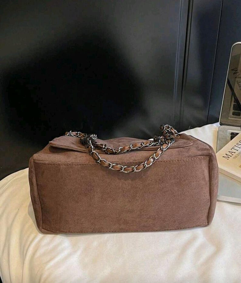 May include: A brown suede shoulder bag with a silver chain strap. The bag has a rectangular shape with a flap closure. The bag is on a white surface.