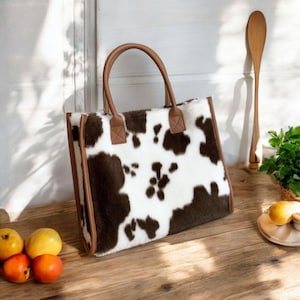 May include: A tote bag with a cowhide pattern in brown and white. The bag has brown handles and trim. The bag is sitting on a wooden surface with fruit and a wooden spoon in the background.