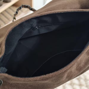 May include: A brown suede handbag with a black interior and a zip closure. The bag has a dark handle and a button closure on the side. The interior features a pocket.