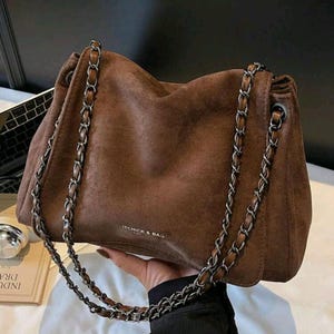 May include: A brown suede handbag with silver chain straps. The bag has a soft, slouchy design and features the text "ITCHICK & BAG" on the front. The chain straps are attached to the top of the bag and hang down on either side.
