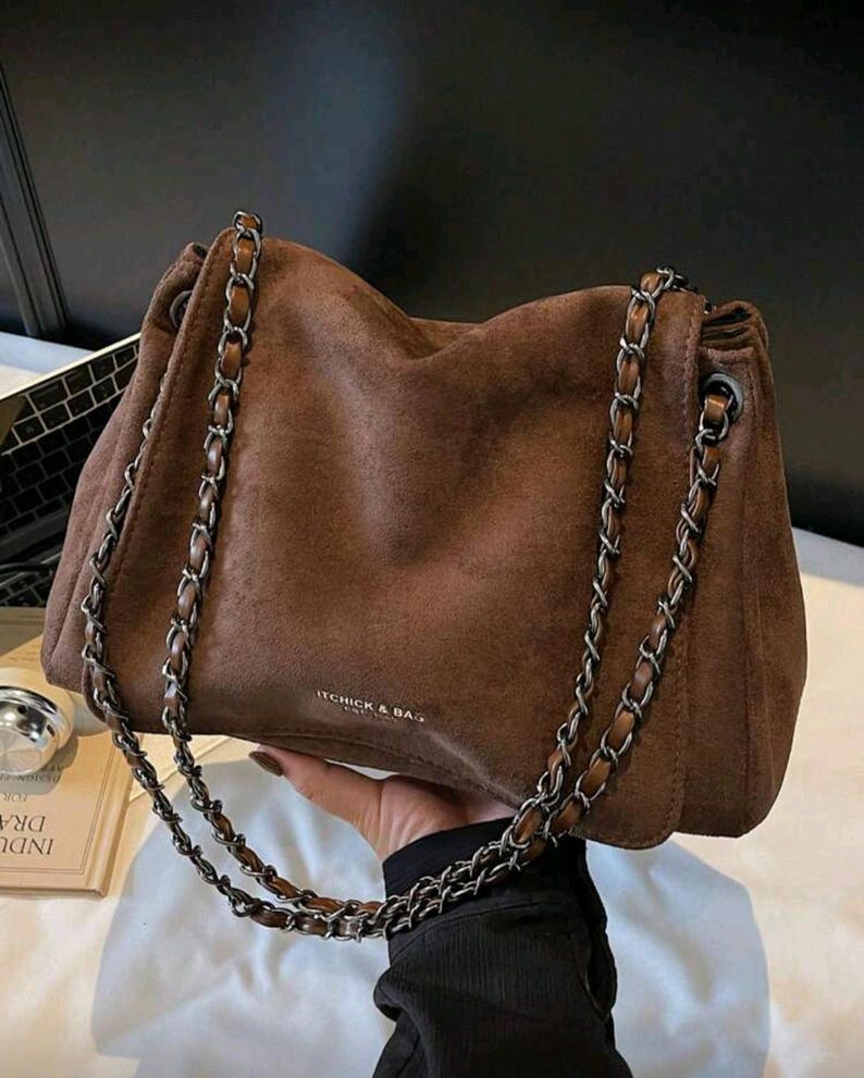 May include: A brown suede handbag with silver chain straps. The bag has a soft, slouchy design and features the text "ITCHICK & BAG" on the front. The chain straps are attached to the top of the bag and hang down on either side.