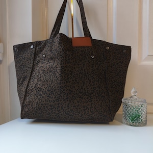 May include: A large tote bag with a brown leopard print pattern. The bag has two handles and a small brown leather patch. The bag is sitting on a white surface.